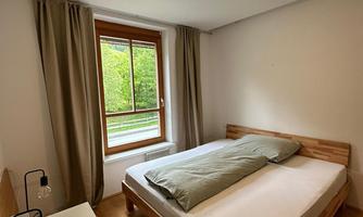 A simple bedroom with a double bed and a large window. The room has light walls and a modern, minimalist style.