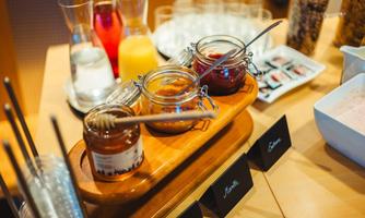 A breakfast table with several small jars containing honey and jam. In the background, drinks and snacks are visible.