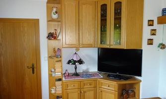 A bright room with a wooden wardrobe and a television. On the wall, there are shelves with various decorative items.