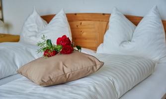 A cozy bed with white pillows and a decorative beige bedding. On the bedding lies a bouquet of red roses and fresh greenery.