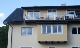 A modern residential building with a balcony and several windows. On the balcony, there are chairs and plants.