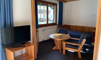 A cozy living space with a table, chairs, and a television. Large windows provide plenty of daylight.