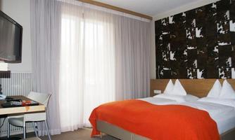 A modern hotel room with a double bed and an orange bedspread. Bright curtains hang at the window, and there is a desk with a television.