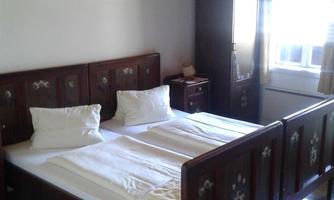 A simple bedroom with a double bed and wooden decor. The room has a wardrobe and windows that provide natural light.