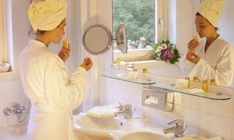 A woman in a bathrobe with a towel around her head stands in front of a mirror. She is applying skincare products and enjoying a moment of relaxation.