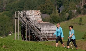 Two women are walking on a hiking trail in a green environment. In the background, a large water wheel can be seen.