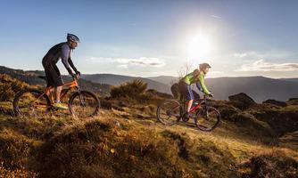 Two mountain bikers are riding on a hilly terrain at sunset. The landscape is characterized by gentle hills and shrubs.