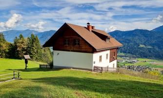 A picturesque house on a green meadow, surrounded by mountains. The sky is blue with white clouds.
