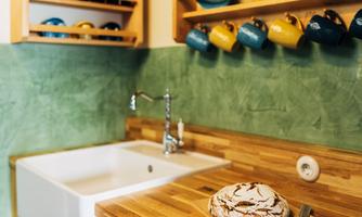 A cozy kitchen with wooden cabinets and green wall paint. On the countertop sits a baked cake, and dishes are neatly hung on a shelf.