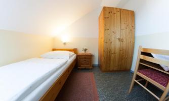 A simply furnished room with a single bed, a nightstand, and a wooden wardrobe. A chair stands next to the bed and the walls are painted light.