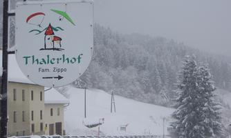 A sign with the name "Thalerhof" and the family name "Zippo" in a snow-covered landscape. In the background, snow-covered trees and a wintry sky can be seen.