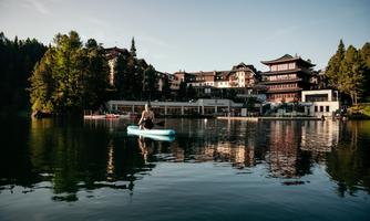 A calm lake with a man on a paddleboard. In the background, a charming hotel and green trees can be seen.