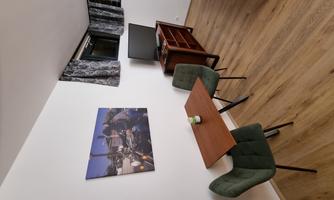 A small, modern living or dining room with a wooden table and two green chairs. A picture hangs on the wall, and there is a television on a wooden piece of furniture.