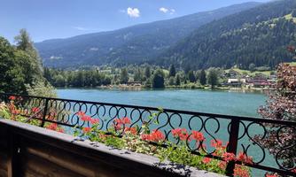 A picturesque view of a clear lake, surrounded by mountains and trees. In the foreground, red flowers bloom on a wooden terrace.