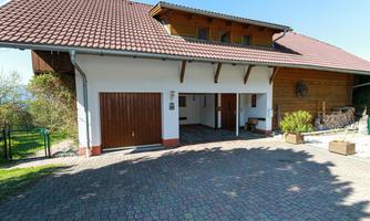 A large house with a red tiled roof and a white facade. In front of the house is a paved driveway and a garage.