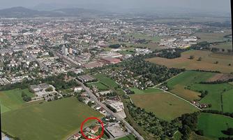 An aerial view of a city with adjacent fields and roads. In the foreground, an area with buildings is marked with a red circle.
