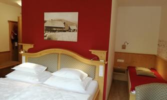 A cozy bedroom with a double bed and a red wall. In the background, there is an additional bed and a simple piece of furniture.