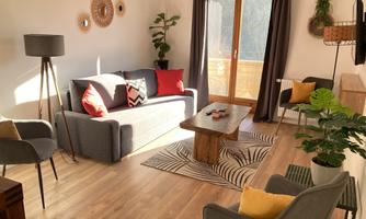 A cozy living room with a gray sofa and colorful cushions. Large windows let in plenty of light, and a wooden table adds a warm atmosphere to the space.