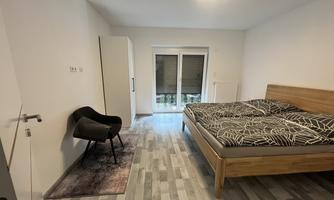 A modern bedroom with a large bed and a cozy armchair. The room has large windows that let in plenty of daylight.