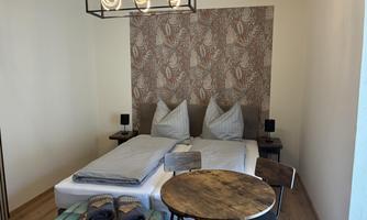 A cozy bedroom with a double bed and a modern lamp. A round wooden table and chairs complement the decor, as well as a balcony with a rattan set.