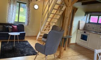 A cozy living area with a sofa, a table, and an armchair. The kitchen is well-equipped, and a staircase leads to the upper area.