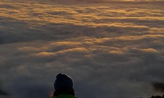 A person is sitting on a mountain, watching the sunrise over a blanket of clouds. The scenery features a snowy landscape and warm tones in the sky.