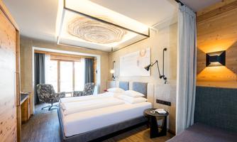 A modern hotel room with two single beds and a cozy seating area. Large windows let in plenty of daylight and offer a view outside.