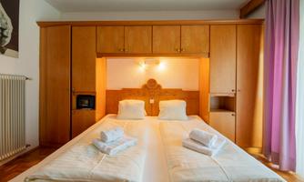 A bright bedroom with a large double bed and wooden cabinets. Fresh towels are laid out on the bed, and a window with purple curtains lets in light.