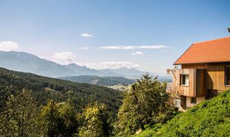 A house on a hill overlooking a picturesque mountain landscape. The sky is clear and sunny, surrounded by green forests.