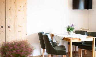 A cozy dining area with a wooden table and green chairs. A television is mounted on the wall and a flower is on the table.