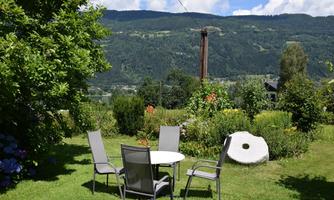 A beautiful garden with a table and chairs, surrounded by green plants. In the background, there are mountains and a blue sky.