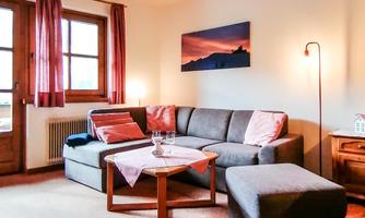 A cozy living room with a gray sofa and a low table. Large windows provide daylight and a warm atmosphere.