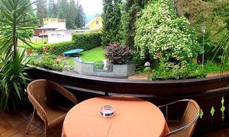 A cozy balcony with a round table and two chairs. In the background, green plants and a beautiful garden are in bloom.