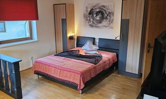 A cozy bedroom with a double bed, bright wooden furniture, and a large window. The bed linen is in vibrant colors and there is a modern artwork on the wall.