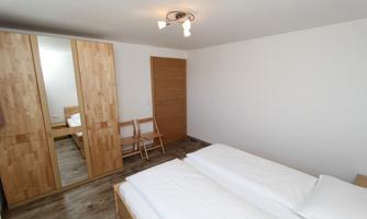 A simple bedroom with two beds and a large wooden wardrobe. There are two chairs and a bright, neutral room.
