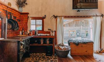 A cozy kitchen with a rustic stove and a window view. The room has a warm wooden floor and simple, natural decorations.