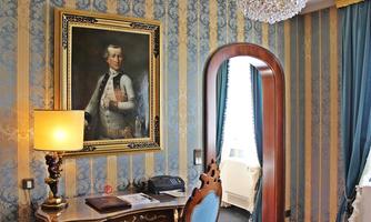 An elegant room with blue wallpaper and a large painting on the wall. An antique desk and a stylish chandelier complement the decor.