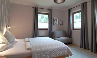 A modern bedroom with a double bed and a cozy armchair. Large windows provide plenty of daylight and a pleasant atmosphere.