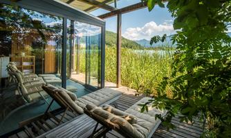 A modern veranda with loungers and a view of a clear lake. Surrounded by lush greenery and mountains in the background.