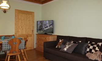 A cozy living room with a sofa, a TV, and a dining table. The walls are painted in a soft green hue and there are wooden details.
