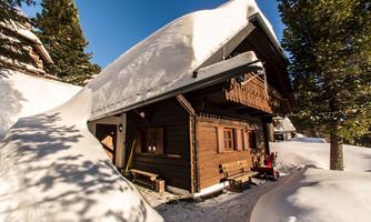 A cozy wooden house with snow on the roof. The surroundings are wintery and idyllic.