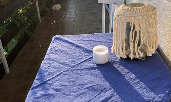 A blue table with a white candle holder and a candle is on a balcony. In the background, you can see plants and a bright room.