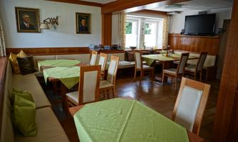 A cozy restaurant with wooden furniture and green tablecloths. Paintings hang on the walls, and there are large windows for natural light.