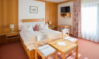 A cozy bedroom with two beds, a small table and chairs. Large windows let in plenty of light and the room is decorated in warm wooden tones.