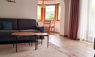 A cozy living room with a gray sofa and a round table. The room has large windows that provide light and a view of the dining area.