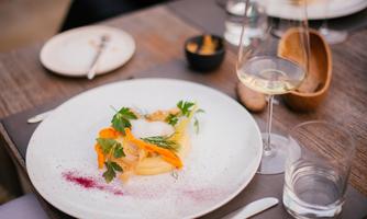 An attractively arranged dish with pasta, fresh herbs, and vegetables. In the background, glasses and plates are visible on a wooden table.