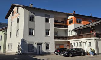 A modern building with multiple floors and a clear blue sky. The facade is partly white and partly orange, surrounded by parked cars.