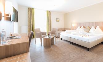 A modern hotel room with a double bed and seating options. The room is bright and cheerfully furnished, with large windows and wooden flooring.