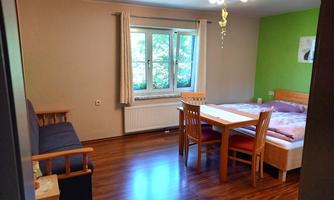 A cozy living room with a table, chairs, and a sleeping area. The walls are painted in soft colors, and large windows provide plenty of light.