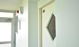 A bright corridor with a white wall and a door with a rectangular window. In the background, an exit can be seen.